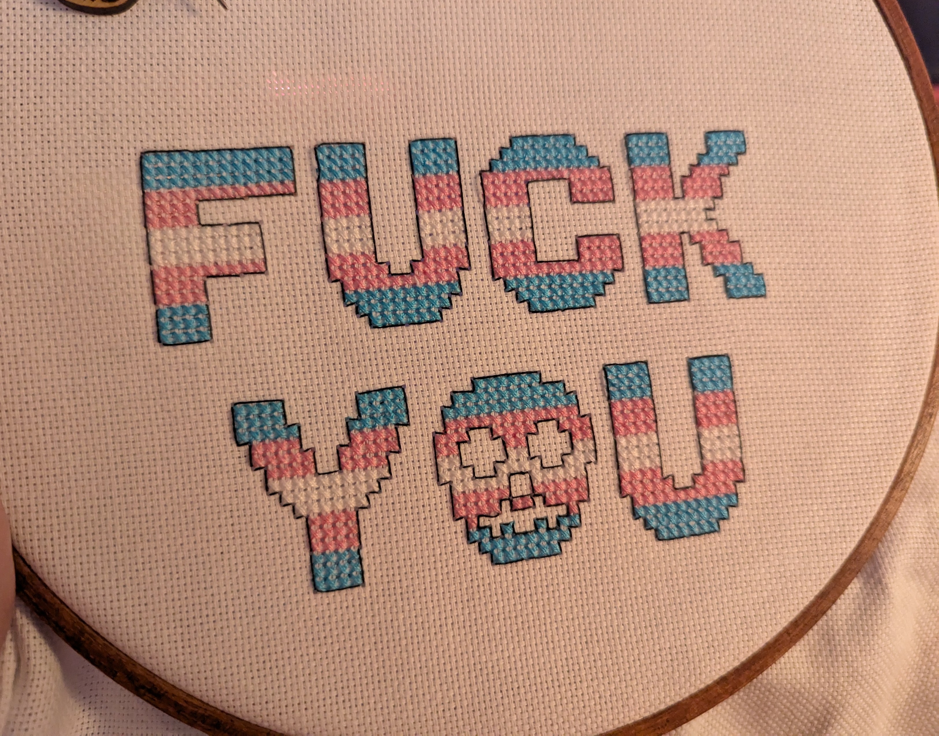 cross stitch project that says FUCK YOU (and the "O" is a skull) in the colors of the transgender flag (blue, pink, white)
