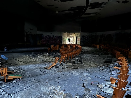 what was once an auditorium for a middle school now lies in ruins. only the orange metal brackets where the chairs were once affixed remain, jutting out of a blue-grey floor littered with shattered glass, metal wiring, broken tile, and other debris of abandonment. only the foreground is well lit; the backdrop is in complete darkness, save for a small corner illuminated by a cell phone flashlight. a woman stands in front of the light source, leaving only her black silhouette visible. 