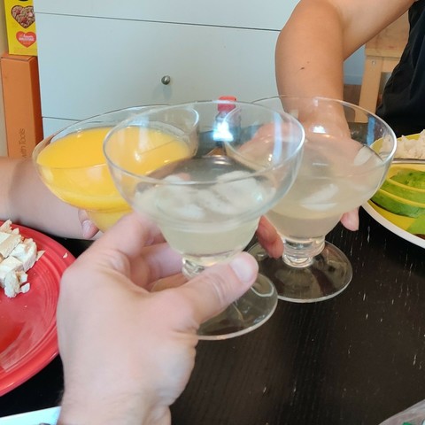 Three margarita glasses clinking together above a kitchen table. Two of them contain margaritas. The one held by a child contains a "margarita".