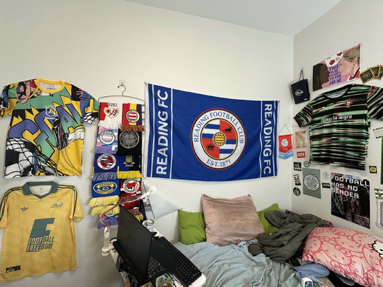 Two walls surrounding my bed. One wall has two football shirts pinned up, on the top a colorful Brazil kit with a graphic of someone kicking a football and the word GOAL, and below it a Bob Marley Jamaica kit that says FOOTBALL IS FREEDOM. Next to that I have several teams' scarves hanging from a pants hanger: Rayo, Celtic, Brøndby, Grorud, Reading, and 3 Clapton scarves. Next to that is a big Reading FC flag.
On the adjoining wall, there is a Portugal training kit with the words "chinga la m…